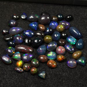May include: A collection of polished black opal gemstones in various shapes and sizes. The opals display a range of colors, including blue, green, red, and orange, with a shimmering, iridescent effect. The gemstones are scattered on a dark surface.