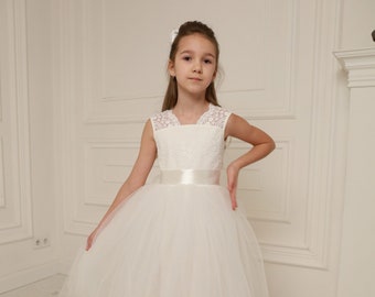 May include: A white tulle dress featuring a lace bodice and a satin ribbon sash. The dress has a full skirt and is knee-length. The girl is wearing white ballet flats and a white bow in her hair. The background is a white room.