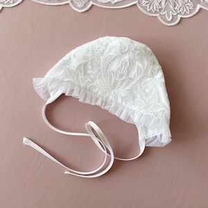 May include: White lace bonnet with a ruffled edge and a white satin ribbon tie. The bonnet is made of delicate lace and has a floral pattern.