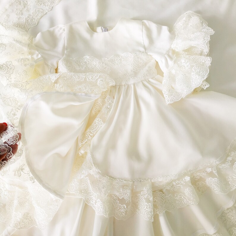 May include: A white satin dress with lace trim and ruffles. The dress has long sleeves and a round neckline. The dress is perfect for a special occasion.