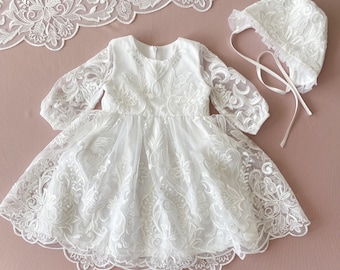Puede incluir: Vestido de bebé blanco de encaje con mangas largas y un gorro a juego. El vestido tiene un delicado patrón floral y un dobladillo festoneado.