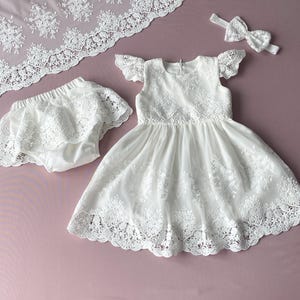 May include: White lace baby dress with a matching lace diaper cover and headband. The dress has a scalloped lace trim and a ruffled neckline.