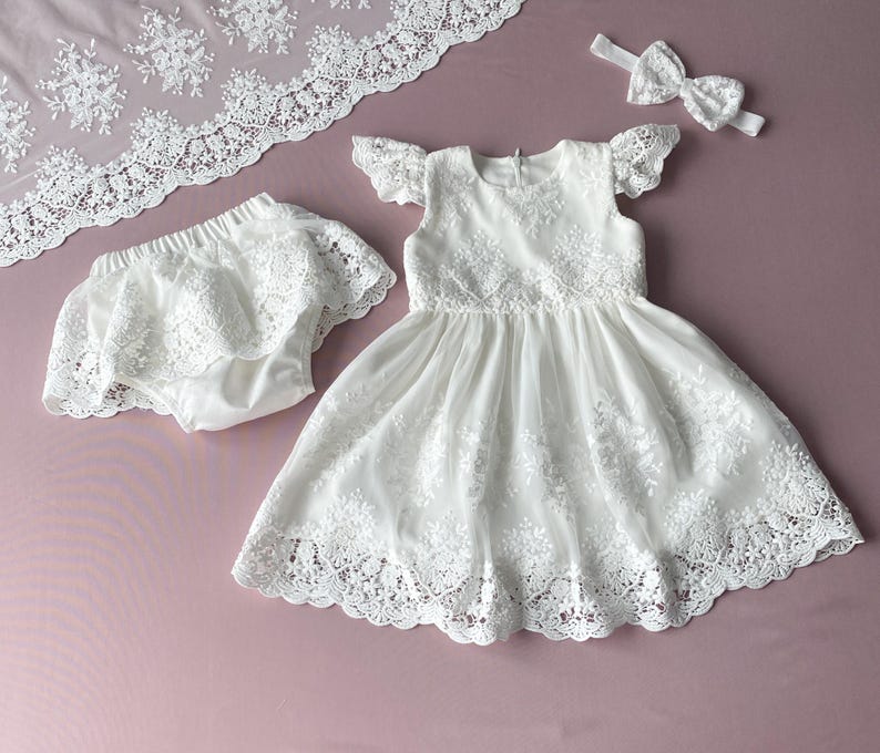 May include: White lace baby dress with a matching lace diaper cover and headband. The dress has a scalloped lace trim and a ruffled neckline.