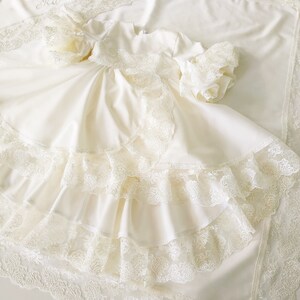 May include: A white baby dress with lace trim and multiple layers of ruffles. The dress has long sleeves and a round neckline.