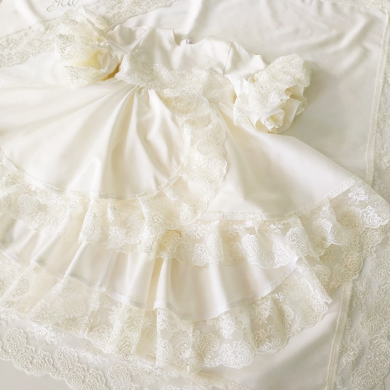 May include: A white baby dress with lace trim and multiple layers of ruffles. The dress has long sleeves and a round neckline.