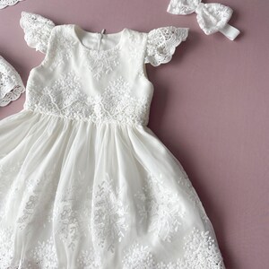 May include: White lace baby dress with a ruffled neckline and short sleeves. The dress has a full skirt and is decorated with delicate lace embroidery. A matching white lace headband is included.