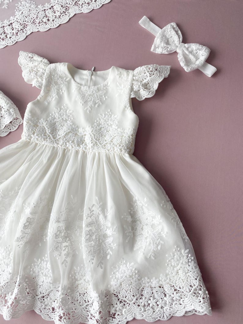 May include: White lace baby dress with a ruffled neckline and short sleeves. The dress has a full skirt and is decorated with delicate lace embroidery. A matching white lace headband is included.