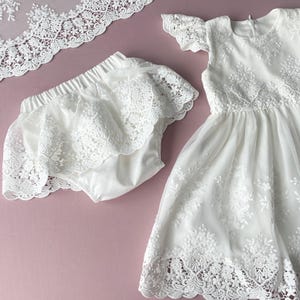May include: White lace dress and matching bloomers for a baby or toddler. The dress has a scalloped lace trim and the bloomers have a ruffled lace trim.