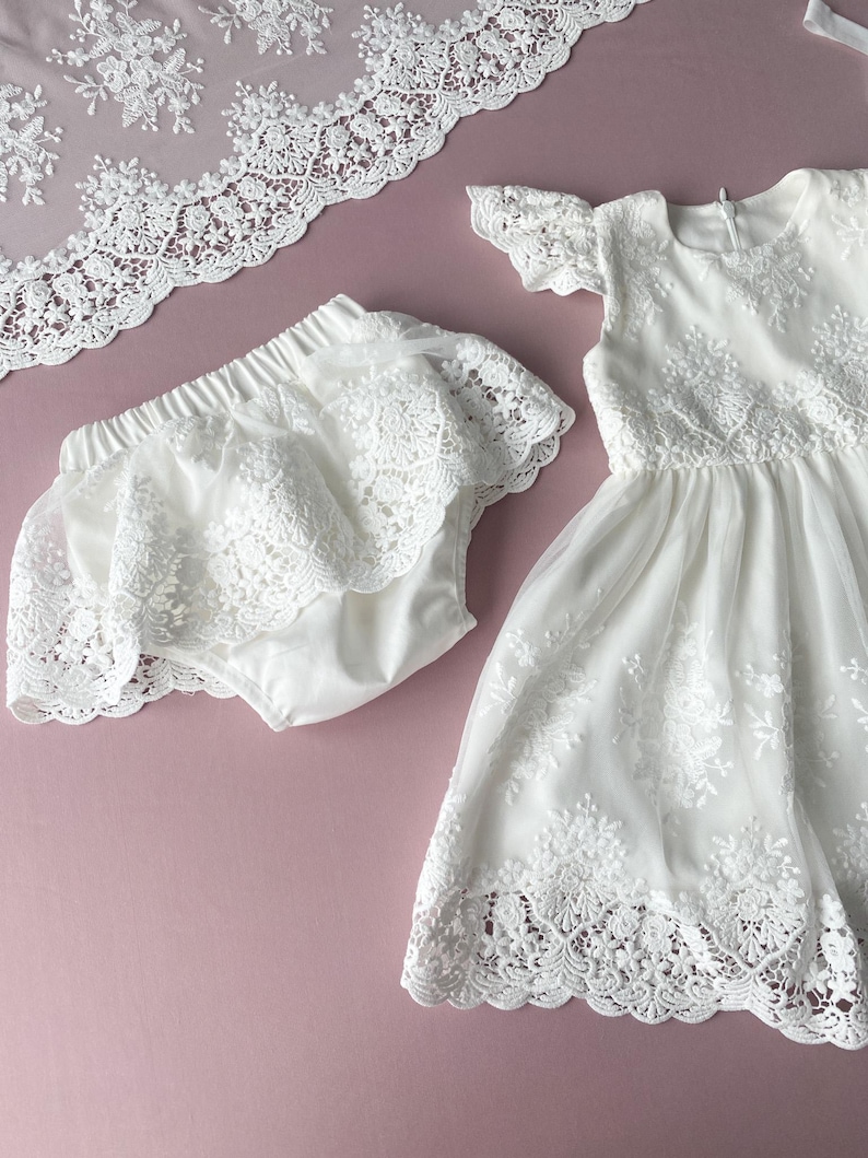 May include: White lace dress and matching bloomers for a baby or toddler. The dress has a scalloped lace trim and the bloomers have a ruffled lace trim.