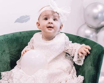 Könnte beinhalten: Ein kleines Mädchen in einem weißen Spitzenkleid mit einem weißen Schleifenhaarband, das einen weißen Ballon hält, während es auf einem grünen Samtstuhl sitzt.