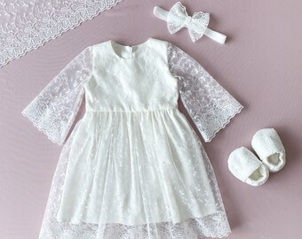 May include: White lace baby dress with long sleeves and a bow headband. The dress has a delicate floral pattern and a full skirt. The headband is tied with a bow in the back.