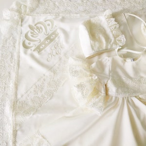 May include: A white satin baby blanket with a lace trim and embroidered crown and initials. The blanket is paired with a matching white lace bonnet and a white satin dress with lace trim.