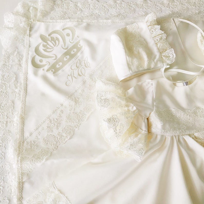 May include: A white satin baby blanket with a lace trim and embroidered crown and initials. The blanket is paired with a matching white lace bonnet and a white satin dress with lace trim.