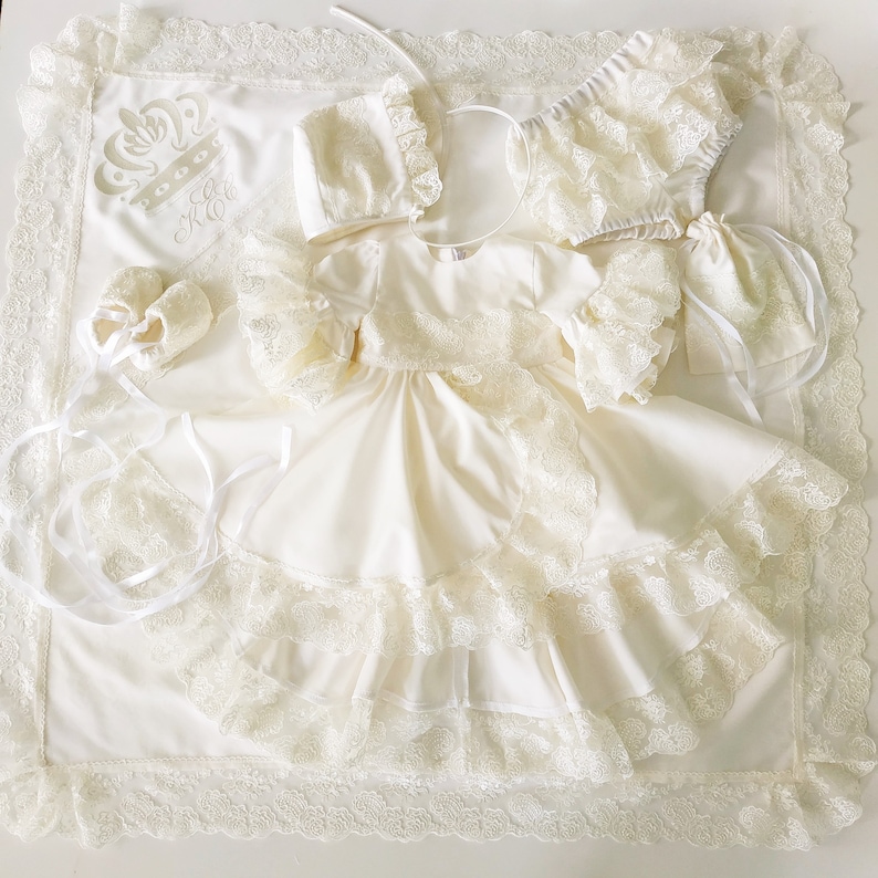 May include: A white baby christening gown with lace trim and a matching bonnet, booties, and diaper cover. The gown has a ruffled skirt and the bonnet has a bow. The set is laid out on a white blanket with a crown and the initials "K.C." embroidered on it.