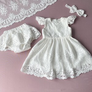 May include: White lace dress with a matching lace diaper cover and headband. The dress has a scalloped lace trim and a ruffled neckline.