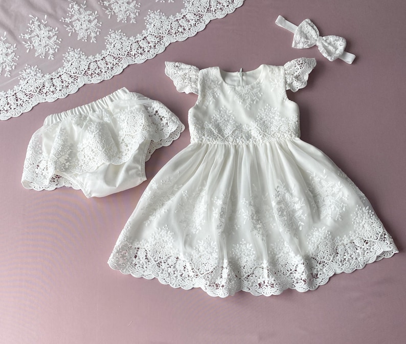 May include: White lace dress with a matching lace diaper cover and headband. The dress has a scalloped lace trim and a ruffled neckline.