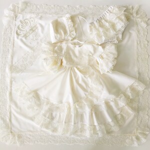 May include: A white baby gown with lace trim and a crown embroidered on the blanket. The gown has a ruffled neckline and sleeves. The blanket has a lace border.