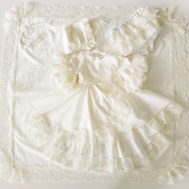 May include: A white baby gown with lace trim and a crown embroidered on the blanket. The gown has a ruffled neckline and sleeves. The blanket has a lace border.