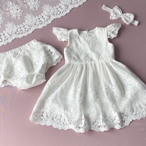 May include: White lace baby dress with a matching lace diaper cover and headband. The dress has a scalloped lace trim and a ruffled skirt.