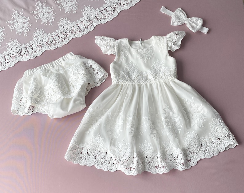 May include: White lace baby dress with a matching lace diaper cover and headband. The dress has a scalloped lace trim and a ruffled skirt.