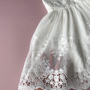 May include: White lace dress with a scalloped hem and floral embroidery. The dress is made of a sheer fabric and has a flowy silhouette.