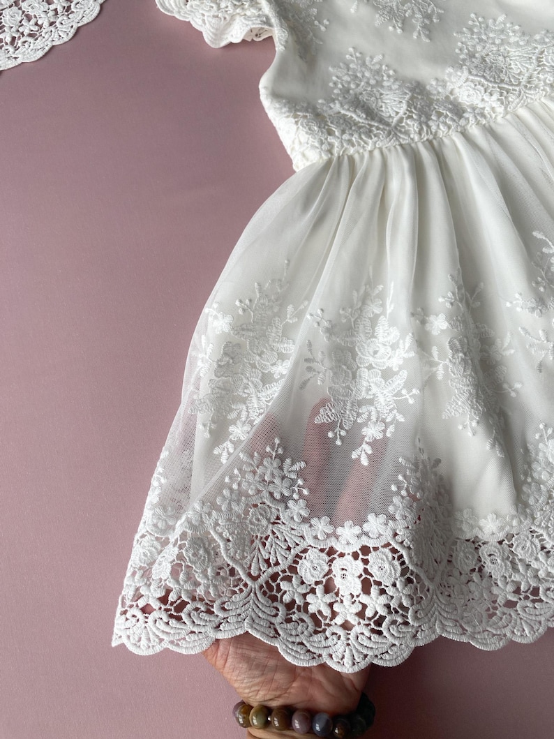 May include: White lace dress with a scalloped hem and floral embroidery. The dress is made of a sheer fabric and has a flowy silhouette.