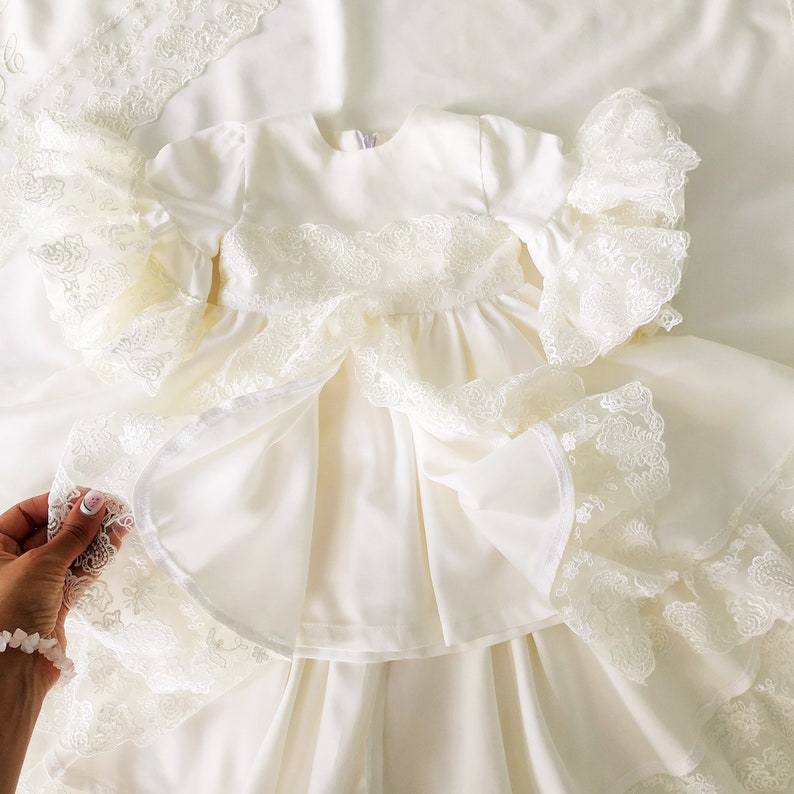 May include: A white baby dress with lace trim and a ruffled skirt. The dress has long sleeves and a round neckline.