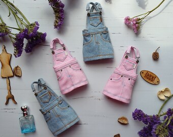 May include: Four miniature denim overalls in two colors: blue and pink. The overalls have a classic design with adjustable straps and pockets. The overalls are arranged on a white wooden surface with purple flowers and other decorative items.
