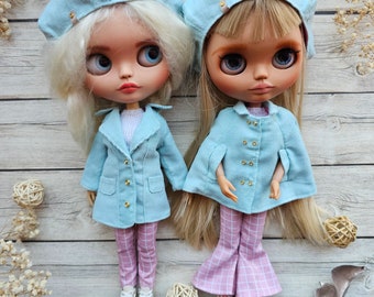 May include: Two dolls with large eyes and long hair wearing matching light blue corduroy berets and jackets. The doll on the left wears a light blue coat over pink plaid pants and white sneakers. The doll on the right wears a light blue cape over pink plaid flared pants and white shoes.