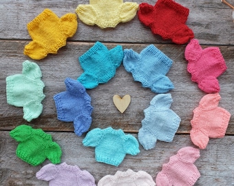 May include: A circle of 13 knitted baby sweaters in various colors, including yellow, red, blue, green, and pink. The sweaters are arranged in a circle on a wooden background.
