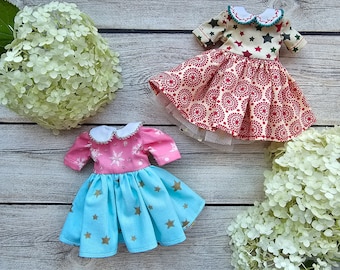 May include: Two miniature doll dresses. One has a pink bodice with snowflake detailing and a light blue skirt with gold stars. The other features a cream top with green and red stars and a red patterned skirt.