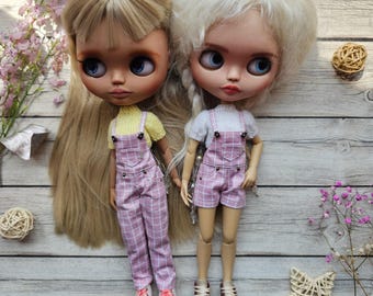 May include: Two dolls with blonde and white hair, wearing matching outfits. One doll has long hair and the other has a braid. Both dolls are wearing pink plaid overalls with white tops and pink shoes. The dolls are standing on a light wood surface.
