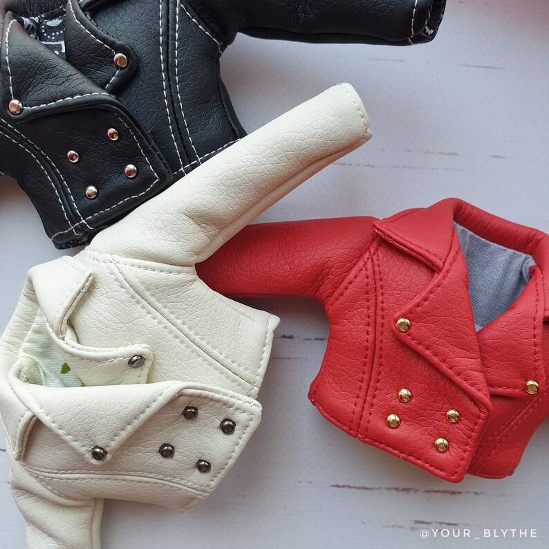 May include: A close-up of three miniature leather jackets in black, cream, and red. The black jacket has silver snaps and white stitching. The cream jacket has silver snaps. The red jacket has gold snaps. All jackets have a classic biker style.