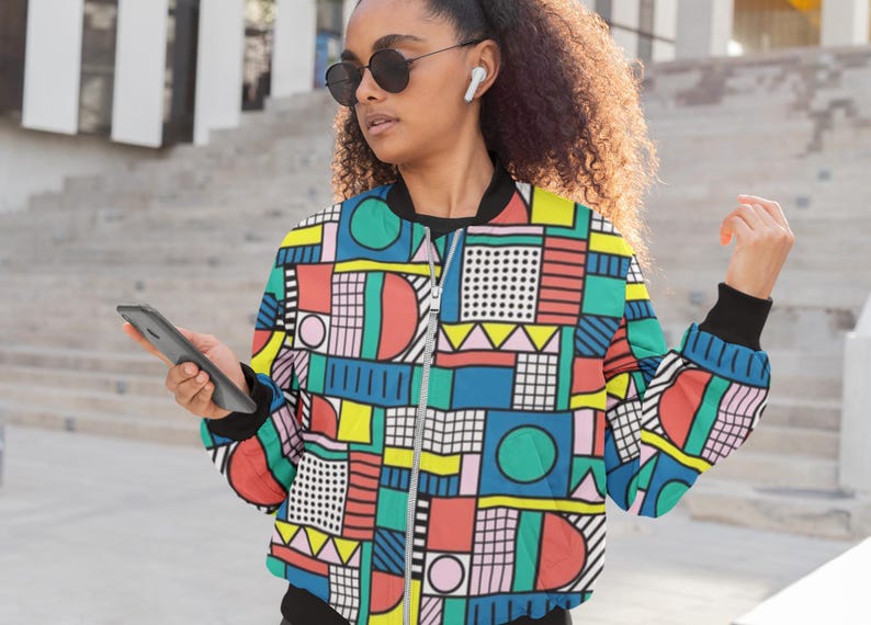 May include: A colorful geometric patterned bomber jacket with a zipper closure. The jacket has a mix of red, blue, green, yellow, pink, and black geometric shapes.