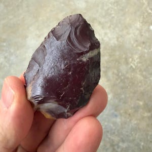 Peut inclure: Un outil en pierre en forme de goutte d'eau, de couleur marron foncé, avec une surface lisse et brillante et des bords écaillés visibles. L'objet présente une pointe effilée et une base arrondie, avec des bandes plus claires près du bas.