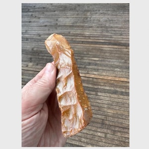 May include: A hand-held, light brown stone tool with a textured surface. The tool has a curved shape and appears to be a primitive cutting or scraping implement. The stone has a natural, earthy color and a rough, uneven texture.