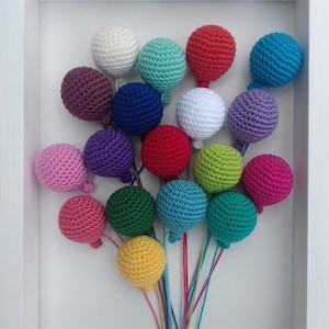 Puede incluir: Una exposición enmarcada de globos de crochet en varios colores, incluyendo rosa, morado, azul, verde, amarillo, rojo y blanco. Los globos están dispuestos en un racimo y parecen flotar hacia arriba.