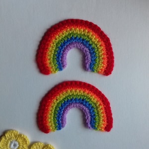 May include: Two crocheted rainbows in red, orange, yellow, green, blue, purple and pink. Two yellow crocheted flowers are below the rainbows.