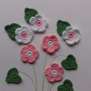 May include: A collection of crocheted flowers and leaves in pink, white, and green. The flowers have a white center and are arranged on a white background.