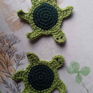 Può includere: Due ornamenti di tartaruga verde all'uncinetto con occhi di perline nere. Le tartarughe hanno un centro verde scuro e bordi verde chiaro.