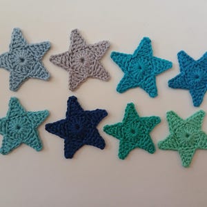 Op de afbeelding: Acht gehaakte stervormen in verschillende tinten blauw en grijs. De sterren zijn allemaal even groot en hebben een vijfpuntig ontwerp.