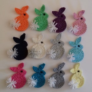 Puede incluir: Doce formas de conejo de crochet en varios colores, incluyendo naranja, verde, morado, rosa, negro, blanco, gris, azul y amarillo. Cada conejo tiene una cola de pompón blanco.