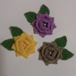 May include: Three crocheted flowers with green leaves. The flowers are purple, yellow, and brown. Each flower has a pearl bead in the center.