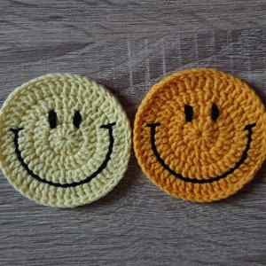 Peut inclure: Deux dessous de verre smiley crochetés. L'un est de couleur jaune clair et l'autre orange doré. Les deux ont des yeux cousus noirs et un sourire courbé. Les dessous de verre sont circulaires et reposent sur une surface en bois.