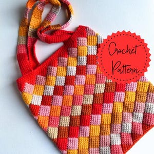 Può includere: Borsa all'uncinetto fatta a mano con un motivo a scacchiera nei toni del rosso, arancione, giallo, rosa e bianco. La borsa ha una lunga tracolla nei colori coordinati e il testo "Crochet Pattern" su un cerchio rosso.