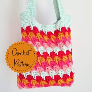 Op de afbeelding: Een handgemaakte gehaakte tote bag met een kleurrijk chevronpatroon. De tas heeft een combinatie van rood, roze, oranje en lichtblauw garen. De tekst "Crochet Pattern" staat op een gouden sticker.