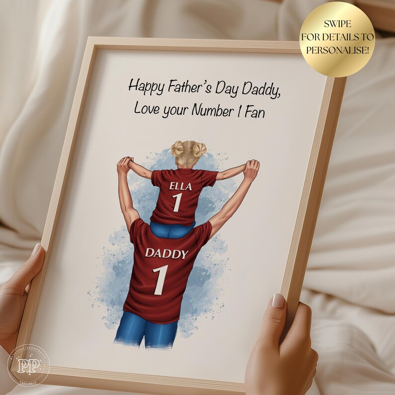 Puede incluir: Una impresi&oacute;n enmarcada con un marco de madera clara. La obra de arte representa a una persona que lleva a un ni&ntilde;o sobre sus hombros, ambos con camisetas granates con el n&uacute;mero 1. El texto dice "Happy Father's Day Daddy, Love your Number 1 Fan."