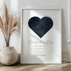 May include: A framed print with a navy blue heart-shaped constellation map on a white background. The text "Our Wedding Day" is displayed, along with names, a date, and the phrase "Forever & Always". A decorative vase with tall, dried plants is in the foreground.