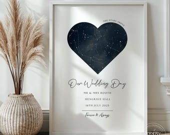 May include: A framed print with a navy blue heart-shaped constellation map on a white background. The text "Our Wedding Day" is displayed, along with names, a date, and the phrase "Forever & Always". A decorative vase with tall, dried plants is in the foreground.