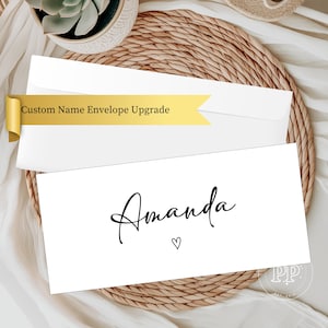 May include: A white card with the name "Amanda" in elegant black script and a small heart symbol. The card is part of a stationery set, with a matching white envelope and a gold banner that reads "Custom Name Envelope Upgrade".